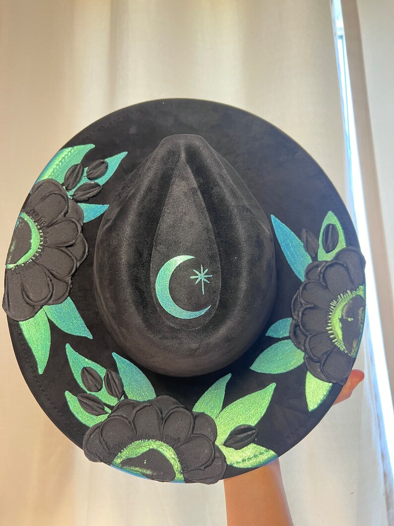 May include: Black wide-brimmed hat with a green and blue floral design. The hat has a crescent moon and star design on the crown.