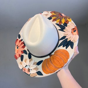K&ouml;nnte beinhalten: Ein wei&szlig;er Filzhut im Fedora-Stil mit breiter Krempe, verziert mit gemalten Herbstblumen, Bl&auml;ttern und einem K&uuml;rbis. Der Hut hat ein schwarzes Band und wei&szlig;e Perlenakzente.