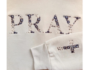 Puede incluir: Sudadera color crema con la palabra "PRAY" bordada en el pecho con un diseño floral. Las letras están rellenas de flores blancas y azul marino. Una cruz floral está bordada en la manga.