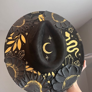 May include: A black felt hat with a wide brim, decorated with gold painted sunflowers, leaves, a crescent moon, a snake, and crystals.