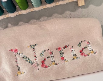 May include: A beige sweatshirt with the word "Nana" embroidered in colorful floral lettering.