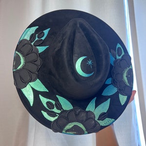 May include: A black felt hat with a wide brim, decorated with teal green floral designs and a crescent moon with a star.