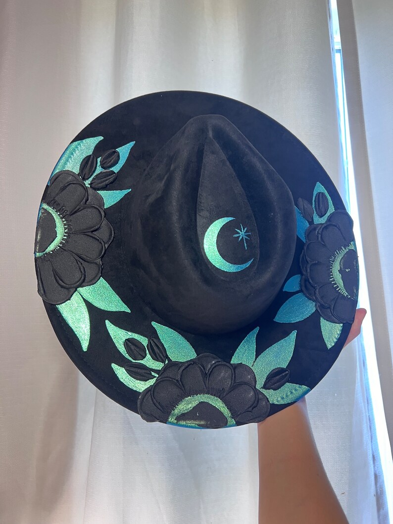 May include: A black felt hat with a wide brim, decorated with teal green floral designs and a crescent moon with a star.