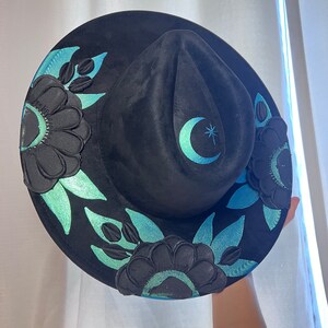 May include: Black felt hat with a crescent moon and star design. The hat is decorated with black floral embroidery and teal accents.