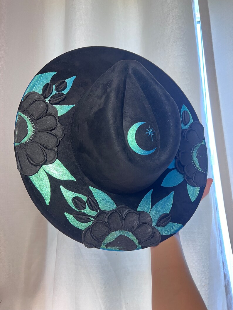 May include: Black felt hat with a crescent moon and star design. The hat is decorated with black floral embroidery and teal accents.