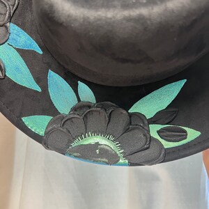 May include: Black hat with a green and blue floral design. The hat has a wide brim and is decorated with a floral pattern.