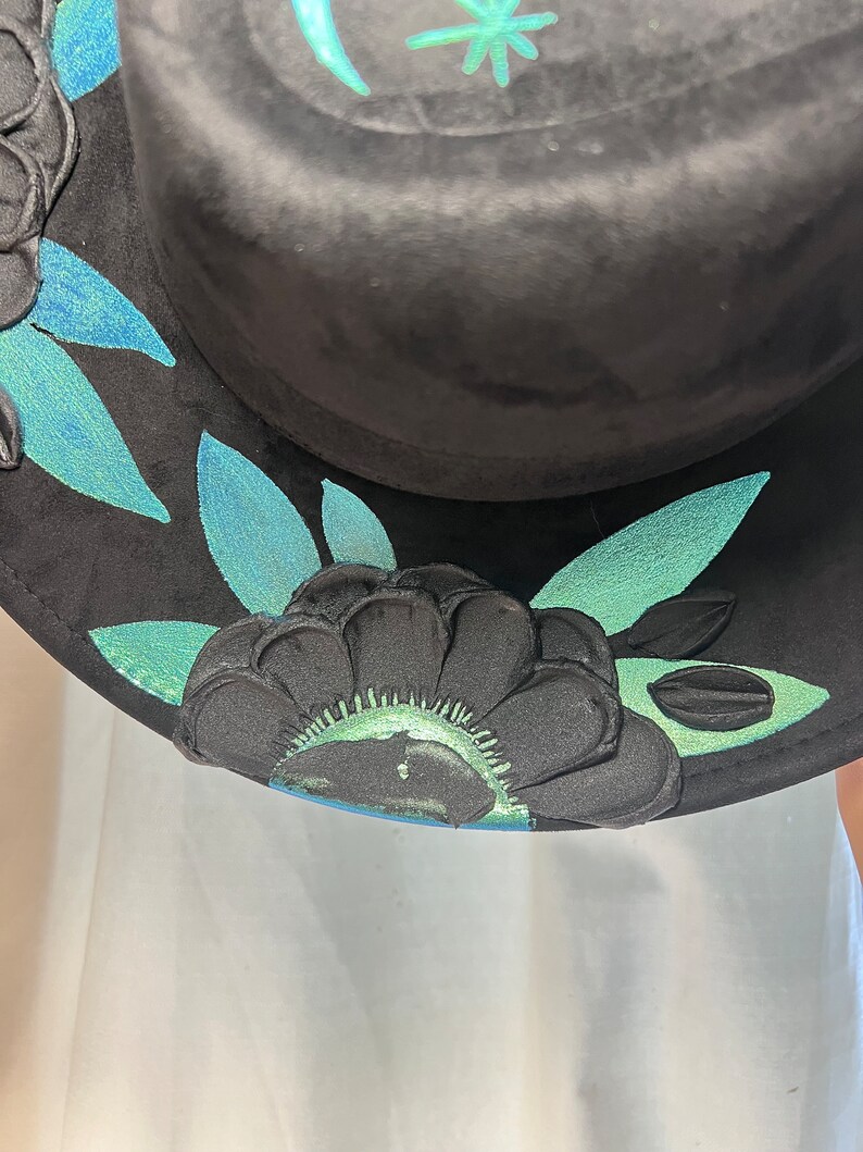 May include: Black hat with a green and blue floral design. The hat has a wide brim and is decorated with a floral pattern.