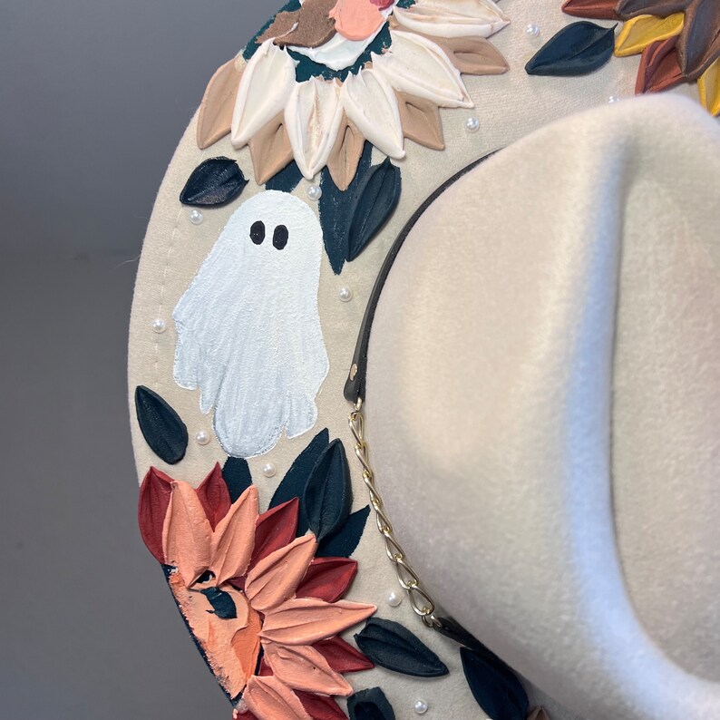 K&ouml;nnte beinhalten: Ein wei&szlig;er Filzhut im Cowboy-Stil mit schwarzem Band und goldener Kette. Der Hut ist mit gemalten Blumen, Bl&auml;ttern und einem Geist verziert.