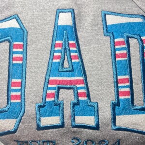 以下が含まれることがあります： グレーのトレーナーに、青と白のストライプで「DAD」が刺繍されています。 「DAD」の下には「EST 2024」と刺繍されています。