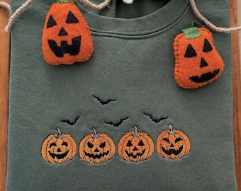May include: An olive green crewneck sweatshirt featuring an embroidered row of four orange jack-o'-lanterns with black facial features and small black bats above them. Two felt pumpkin ornaments and a white felt ghost are attached by twine.