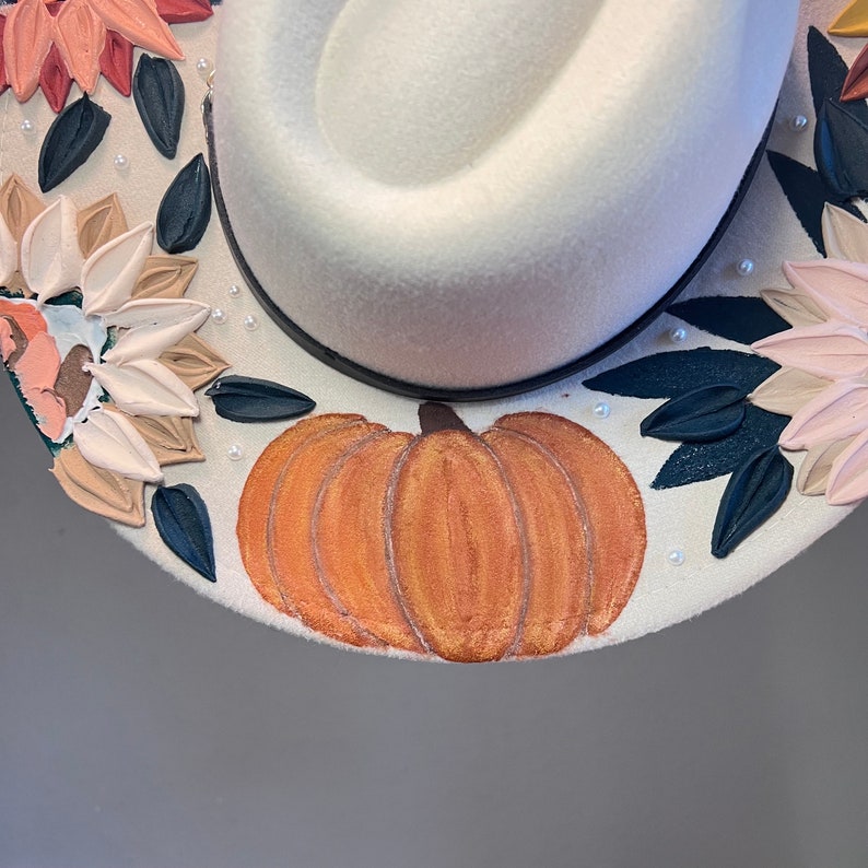 K&ouml;nnte beinhalten: Ein wei&szlig;er Filzhut mit einem bemalten K&uuml;rbisdesign. Der Hut ist mit Blumen und Bl&auml;ttern in Rosa-, Wei&szlig;- und Gr&uuml;nt&ouml;nen verziert. Der K&uuml;rbis ist in Orange- und Braunt&ouml;nen bemalt.