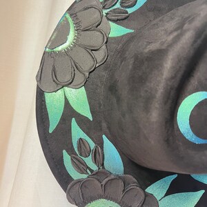 May include: Black witch hat with green and blue floral embroidery. The hat has a wide brim and a tall crown.