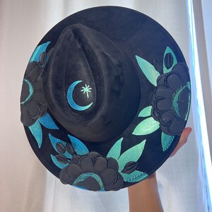 May include: Black velvet hat with a crescent moon and star design embroidered in teal and white. The hat is decorated with teal and black floral embroidery.