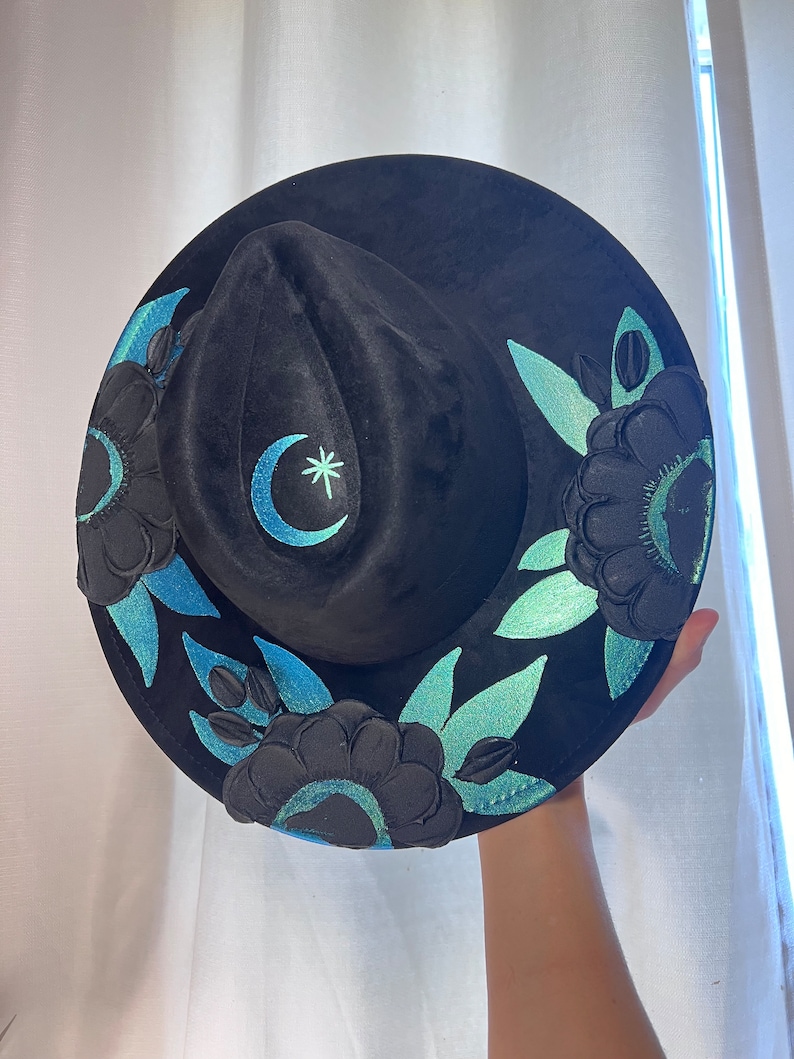 May include: Black velvet hat with a crescent moon and star design embroidered in teal and white. The hat is decorated with teal and black floral embroidery.