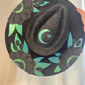 May include: A black velvet hat with a crescent moon and star design. The hat is decorated with green and black floral embellishments.