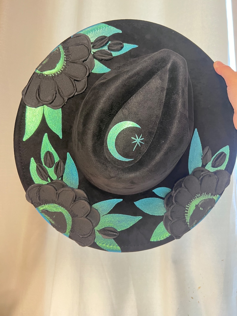 May include: A black velvet hat with a crescent moon and star design. The hat is decorated with green and black floral embellishments.