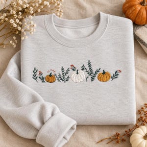 May include: A light gray crewneck sweatshirt featuring an embroidered design across the chest. The embroidery depicts three pumpkins in orange and white, surrounded by green leafy branches and small red berries, creating a seasonal botanical pattern.