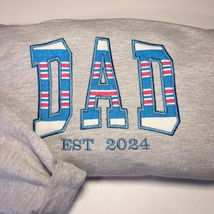 以下が含まれることがあります： グレーのトレーナーに、青、白、赤のストライプで「DAD」という文字が刺繍されています。「EST 2024」という文字が「DAD」の下に刺繍されています。