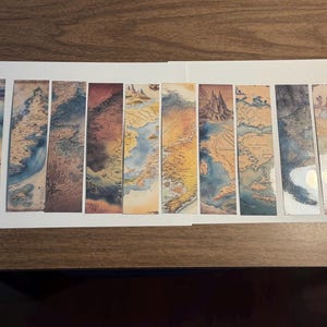 May include: A collection of ten rectangular bookmarks featuring antique map designs. The bookmarks display various color palettes, including blues, browns, and yellows, with detailed illustrations of landmasses, oceans, and compasses. The bookmarks are arranged on a white surface.