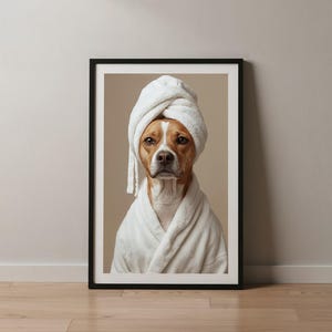 Puede incluir: Una impresi&oacute;n art&iacute;stica enmarcada que muestra a un perro de color canela y blanco con un albornoz de felpa blanco y una toalla blanca a juego envuelta alrededor de la cabeza. El retrato tiene un fondo beige neutro y un marco negro.