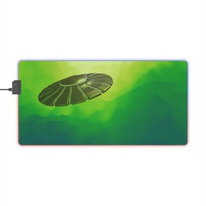 Op de afbeelding: Een groene en blauwe muismat met een zilveren UFO die over een groene en blauwe nevelachtergrond vliegt.