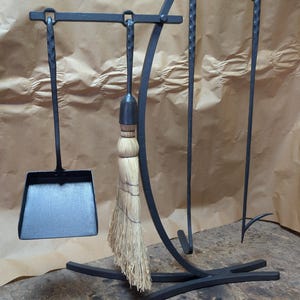 Może przedstawiać: Czarny zestaw narzędzi do kominka z kutego żelaza. Zestaw zawiera łopatę, miotłę, pogrzebacz i stojak. Narzędzia są zawieszone na zakrzywionym stojaku z podstawą w kształcie krzyża.