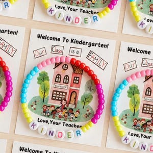 May include: A circular bracelet made of multicolored plastic beads featuring white letter beads that spell out KINDER. The bracelet is displayed on a card with a printed illustration of a schoolhouse and the text Welcome To Kindergarten!