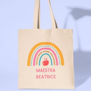 May include: A beige tote bag featuring a rainbow design with the words "Teach Love Inspire Motivate Listen Encourage" surrounding the rainbow. The bag also includes a red apple and the text "Maestra Beatrice" beneath the rainbow.