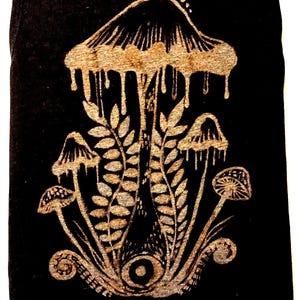 Könnte beinhalten: Ein schwarzes Tanktop mit einem metallisch goldenen Grafikdruck eines großen zentralen Pilzes mit tropfenden Rändern, flankiert von kleineren Pilzen und farnartigem Blattwerk. An der Basis des zentralen Pilzes befindet sich ein kreisförmiges Auge.
