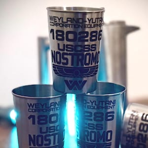 Peut inclure: Verres à shot en métal argenté empilés, avec le texte "WEYLAND-YUTANI CORPORATION EQUIPMENT 180286 USCSS NOSTROMO". Les verres sont cylindriques et ont une finition mate. Le design comprend un logo stylisé.