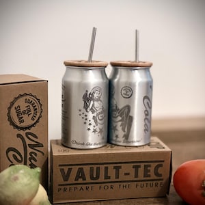 Op de afbeelding: Twee zilverkleurige drankblikjes met bamboe deksels en metalen rietjes, met illustraties in retrostijl en het woord "Cola". De blikjes staan op een bruine doos met de tekst "VAULT-TEC PREPARE FOR THE FUTURE."