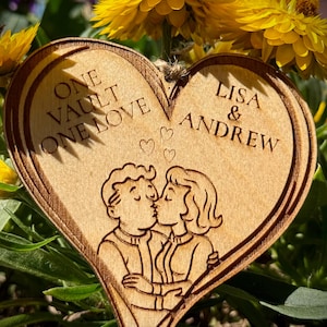 Puede incluir: Un adorno de madera en forma de corazón con el texto grabado "ONE VAULT ONE LOVE" y "LISA & ANDREW". Presenta una ilustración de una pareja besándose. El fondo incluye flores amarillas y follaje verde.