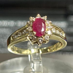 May include: A gold ring featuring a large oval ruby center stone surrounded by smaller diamonds. The ring's band is also adorned with diamonds, creating a halo effect around the ruby. The ring is displayed on a clear stand.