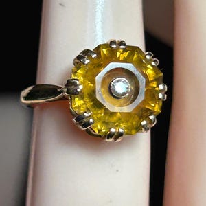 Op de afbeelding: Een gouden ring met een grote, gefacetteerde gele edelsteen en een centrale witte steen. De ring heeft een decoratieve zetting rond de gele steen en de band is van gepolijst goud. Het ontwerp van de ring is opvallend.
