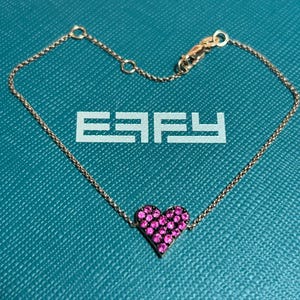 May include: A gold-toned bracelet with a heart-shaped pendant. The heart is encrusted with small, pink gemstones. The bracelet is on a textured, teal surface, with the letters "EAFY" visible.