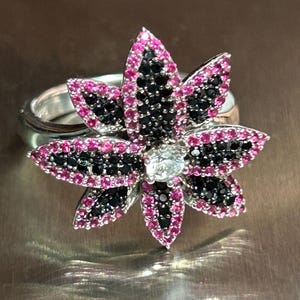 Könnte beinhalten: Ein silberner Ring mit einem Blumenmuster. Die Blume hat Blütenblätter mit rosa und schwarzen Edelsteinen, die einen zentralen klaren Edelstein umgeben. Der Ring ist vor einem metallischen Hintergrund platziert.
