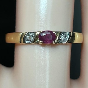 May include: A gold band ring featuring a central oval-cut deep red ruby gemstone flanked by two small clear accent stones set in silver-toned metal settings on either side of the ruby, creating a contrast against the polished gold band.
