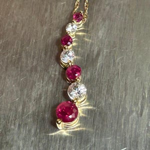 May include: A gold necklace featuring a vertical pendant with a series of alternating round-cut red rubies and clear white diamonds. The stones are set in a graduated arrangement, increasing in size toward the bottom, suspended from a gold chain.