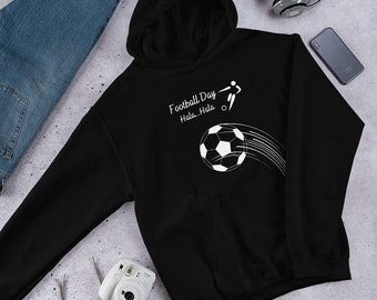 May include: Black hooded sweatshirt with a white graphic of a soccer ball and a player kicking it. The text "Football Day Hala...Hala" is above the graphic. The sweatshirt is on a light gray surface with blue jeans, headphones, and other accessories.