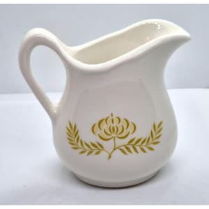 May include: Off-white ceramic creamer with a curved handle and spout. The creamer has a gold floral design on the front, featuring a central flower and leafy accents. A classic piece for serving cream or milk.
