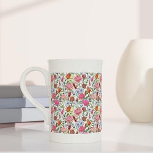 Op de afbeelding: Een witte keramische mok met een bloemenpatroon met roze, rode en blauwe bloemen. De mok heeft een gebogen oor en staat op een wit oppervlak. Vazen en boeken zijn op de achtergrond te zien.