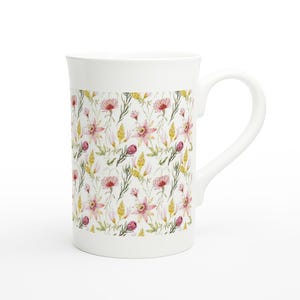 Op de afbeelding: Witte keramische mok met een bloemenpatroon. Het ontwerp toont roze en gele bloemen met groene bladeren op een witte achtergrond. De mok heeft een gebogen handvat.