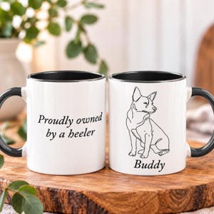 May include: Two white ceramic mugs with black interiors and handles. One mug features the black text "Proudly owned by a heeler". The other mug displays a black line art illustration of a dog and the name "Buddy" printed in black text below it.