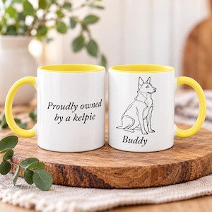 Könnte beinhalten: Zwei weiße Keramiktassen mit leuchtend gelbem Innenraum und Henkel. Eine Tasse zeigt den schwarzen Schriftzug "Proudly owned by a kelpie", die andere eine schwarze Strichzeichnung eines Hundes mit dem darunter gedruckten Namen "Buddy".