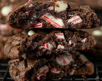 Puede incluir: Una pila de tres galletas de chocolate con bastones de caramelo triturados y chispas de chocolate blanco. Las galletas están en una rejilla de alambre negro.