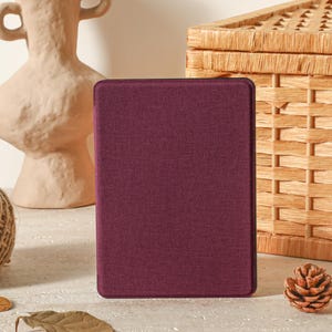 Op de afbeelding: Een bordeauxrode tablethoes met een textuurafwerking staat rechtop. Een decoratieve vaas, een bol touw, een dennenappel en een rieten mand staan op de achtergrond en creëren een natuurlijke, rustieke esthetiek.