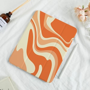 May include: Orange and cream-colored tablet case with a wavy, retro design. The case is open, revealing the screen. A white stylus rests nearby. The case is on a white surface with a brown bottle and flowers.
