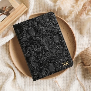 May include: Black leather journal with an embossed floral design. The journal has gold initials "NK" in the bottom right corner. The journal is on a tan plate, with a wooden picture frame and a roll of twine.