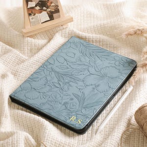 May include: A light blue tablet case with a floral embossed design and black trim. The case has the initials "R.S." in gold. A white stylus and a roll of twine are next to the case. A small wooden easel with a picture is in the background.