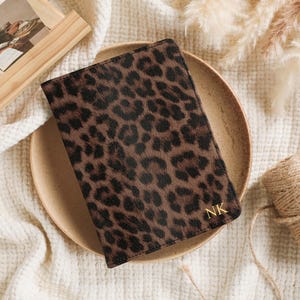 May include: A brown and black leopard print journal with gold initials "NK" on the bottom right corner. The journal is on a tan plate, with a wooden picture frame and a roll of twine in the background.